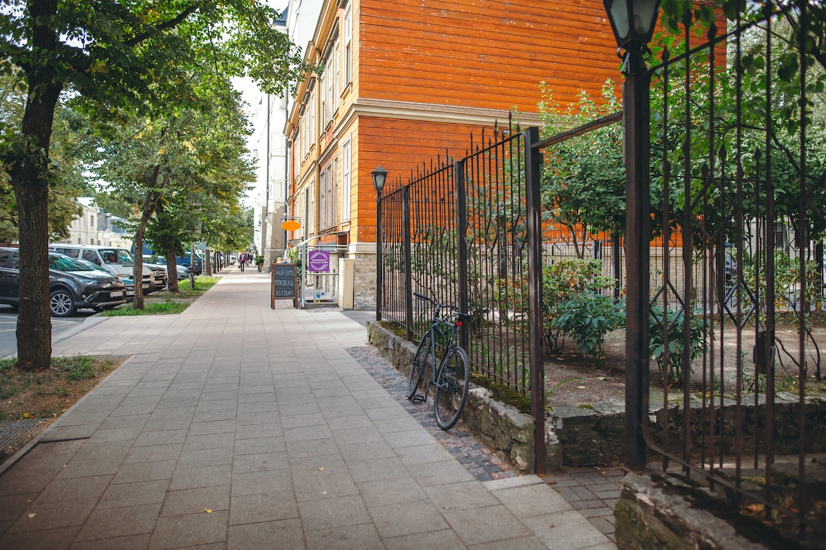A peaceful street in the embassy district features tree-lined sidewalks and a view of an orange wooden building. Bicycle racks and a variety of parked cars are visible, alongside greenery and fences that create a serene atmosphere.