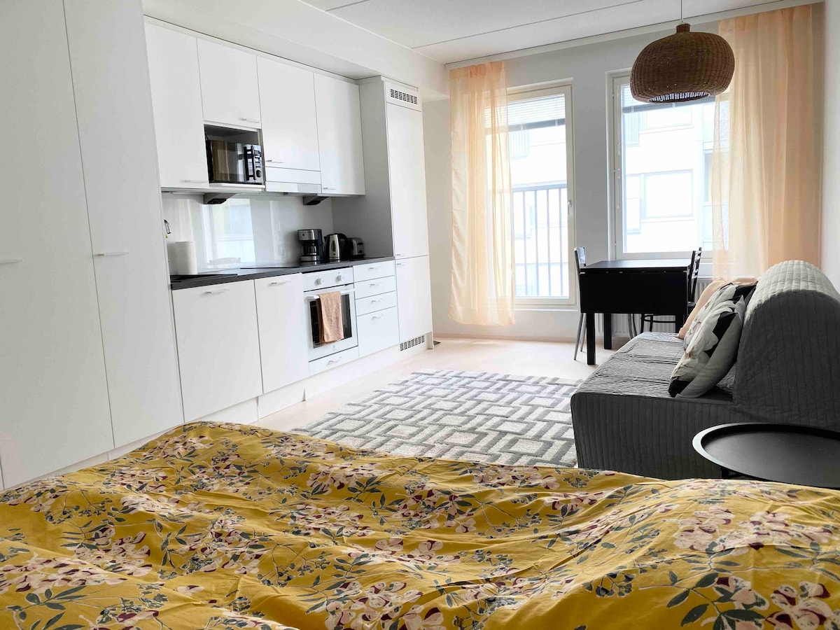 A bright studio space features a well-equipped kitchen with sleek white cabinetry and essential appliances. A comfortable seating area includes a grey couch adjacent to a small dining table. Natural light enters through large windows adorned with light curtains, creating a warm atmosphere.