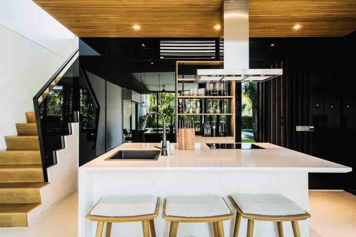 A modern kitchen features a large island with a sleek white countertop and three wooden stools. Understated cabinetry and open shelving showcase an organized space. Natural light enters through large glass openings, illuminating the minimalist design and enhancing the connection to the outside.
