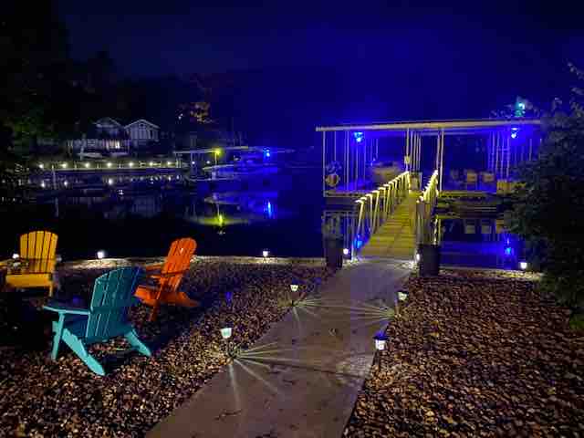 A tranquil lakeside setting is illuminated at night by soft blue lights. A wooden dock extends over calm water, flanked by colorful adirondack chairs in vibrant orange and turquoise. Pathway lights guide the way along a stony shore, creating a serene atmosphere.