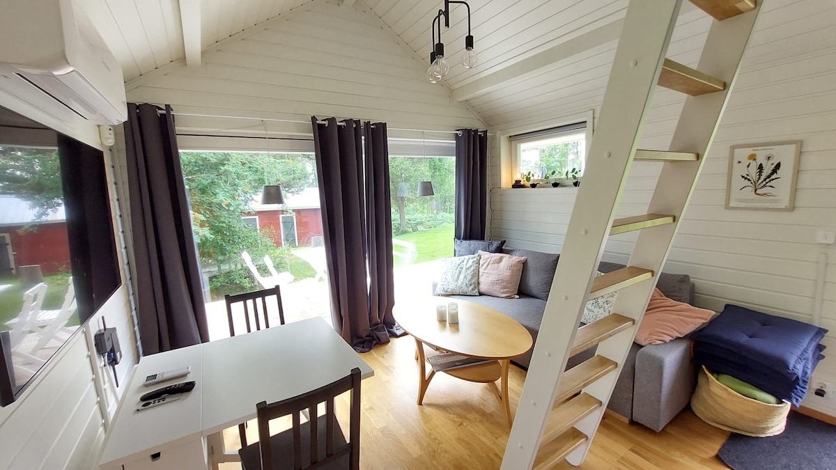 The interior features a bright and airy living area with large windows allowing natural light. A cozy gray sofa is positioned beside a round wooden coffee table, while a work desk is visible. A staircase leads to the sleeping loft, enhancing space utilization.