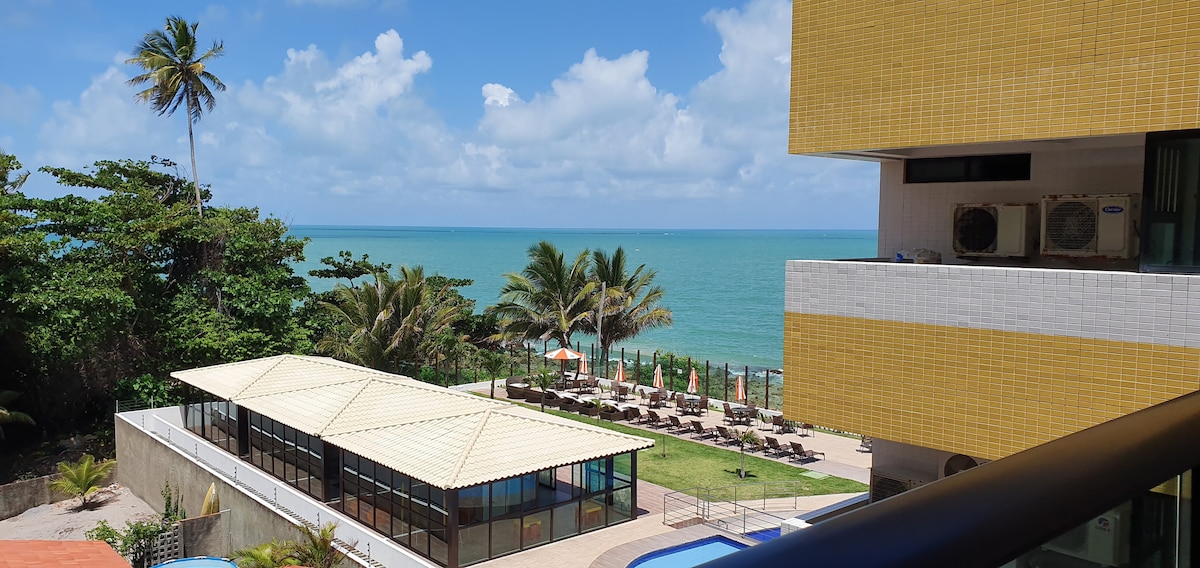 A view of the coastline is framed by lush palm trees under a clear blue sky. Lounge chairs and umbrellas are arranged around a swimming pool. A spacious building with a light-colored roof is visible, offering direct access to the beach.