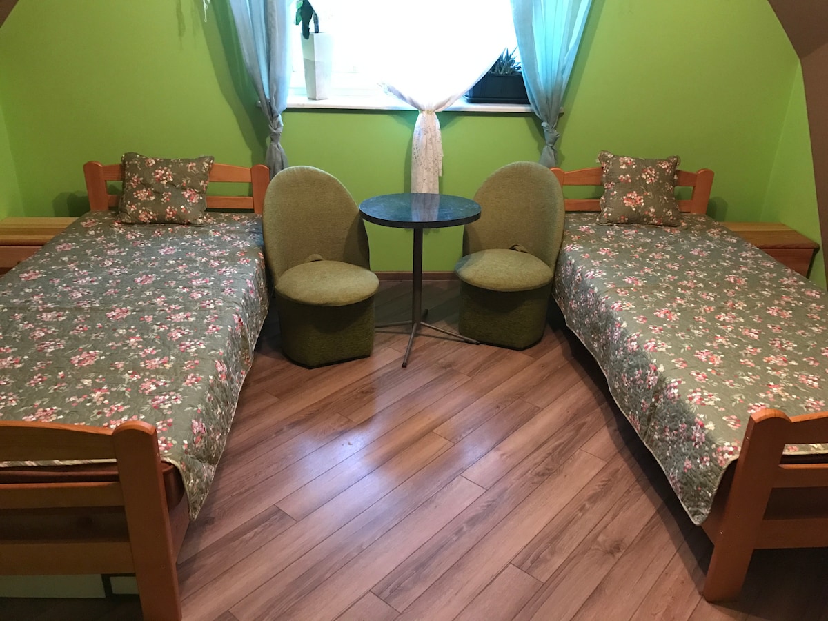 The image features a sleeping area with two wooden beds, each dressed in floral-patterned bedding. A small round table is placed between two green seating chairs. Light enters through a window adorned with sheer curtains, creating a relaxing atmosphere in the space.