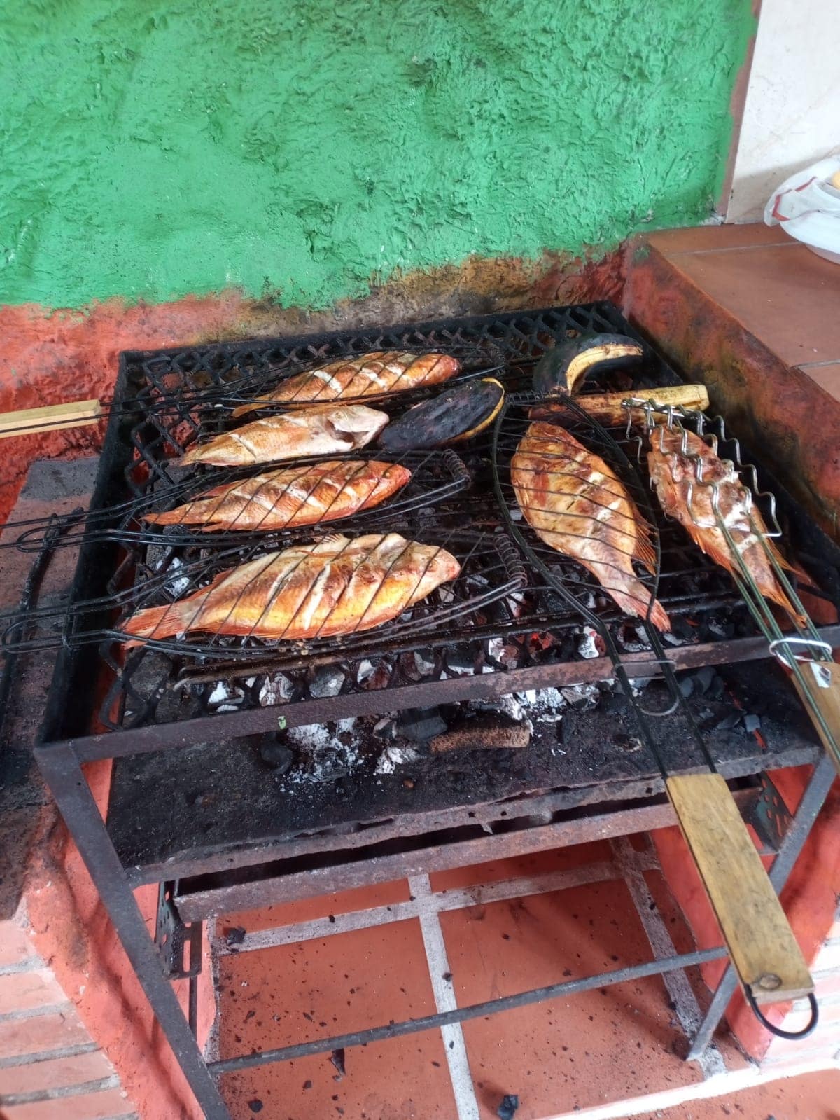 Several fish are being grilled on a charcoal grill, with visible marks from the grilling process. The grill is positioned against a colorful wall, showcasing vibrant green and orange tones. Brick flooring is partially visible in the foreground.