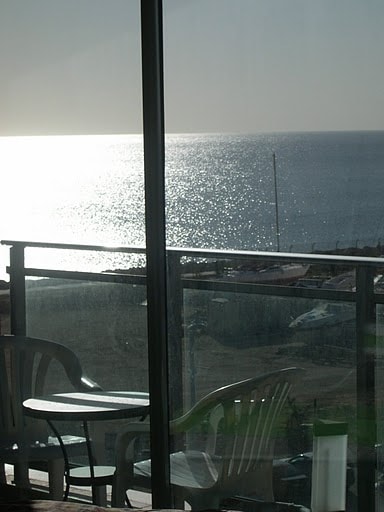 A balcony setting is visible with two white chairs and a small table. The glass balcony door reflects sunlight and offers a view of the shimmering Mediterranean Sea. The horizon is met by a clear sky, creating a serene atmosphere.