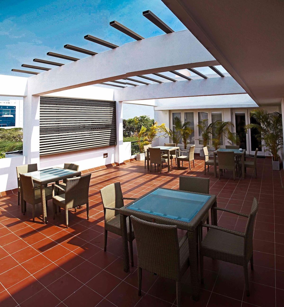 An outdoor terrace is presented with several glass-top tables surrounded by wicker chairs. Lush greenery is visible in the background, while a trellis structure above provides partial shade. The space is designed for relaxation and social gatherings.