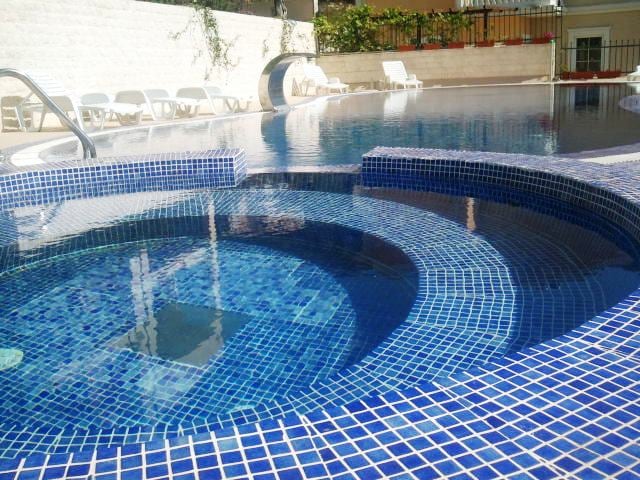 A circular hot tub with a mosaic of blue tiles is positioned beside a tranquil swimming pool. The pool features a gradient of blue hues, reflecting sunlight. White lounge chairs are arranged in the background, providing a relaxing space for guests.