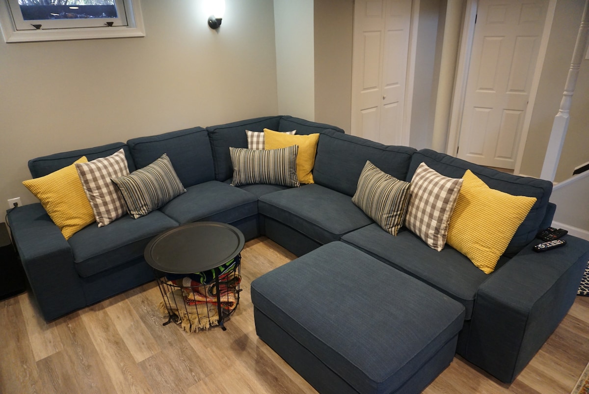 A sectional couch in a neutral tone features an arrangement of yellow and patterned cushions. A small round table is positioned in front, and light-colored wood flooring adds warmth to the living space. Two entrances are visible in the background.