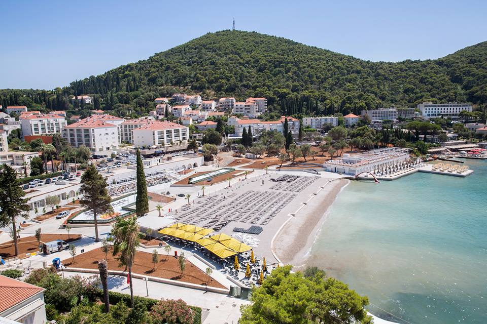 A view of Uvala Beach is presented, showcasing a sandy shoreline meeting clear blue water. Sun loungers are arranged neatly on the beach, with umbrellas providing shade. The backdrop features green hills and a cluster of buildings among greenery, creating a serene coastal atmosphere.