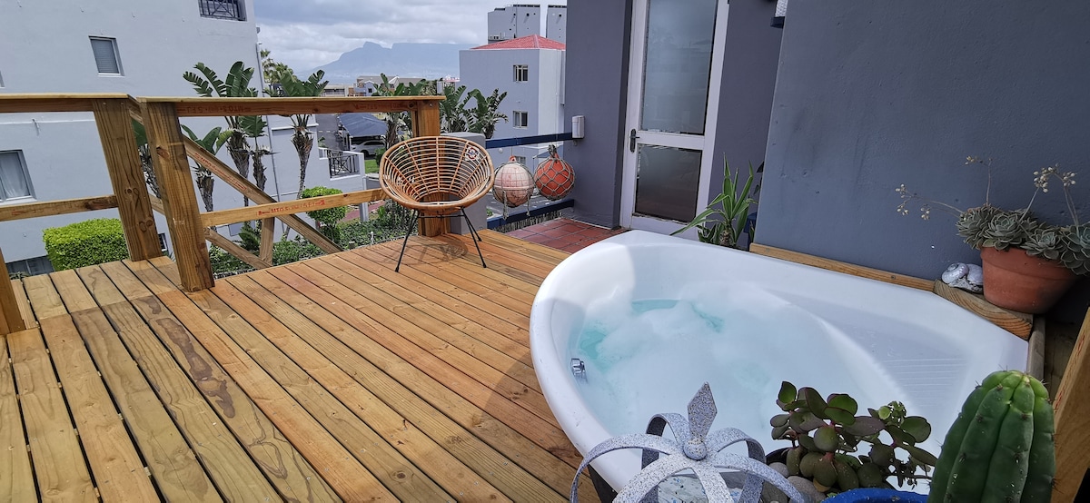 An outdoor deck is featured with a built-in bath and wooden flooring. A simple seating area includes two woven chairs. Lush greenery and potted plants are visible, set against a backdrop with distant views of mountains and buildings.
