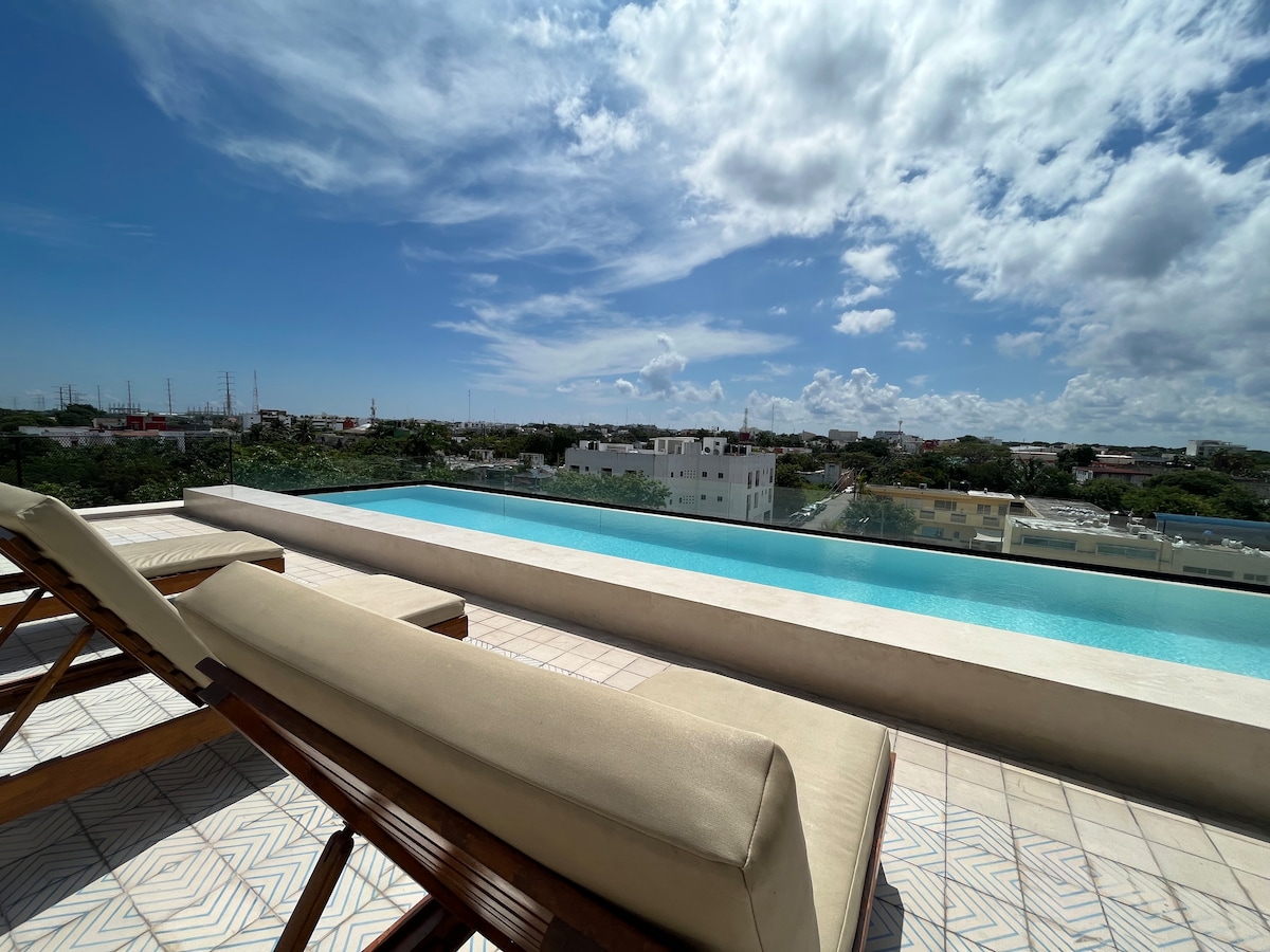 A serene rooftop pool area is visible, featuring a tranquil infinity edge. Two lounge chairs are positioned beside the pool, offering views of the surrounding urban landscape under a partly cloudy sky. The calm water reflects the blue sky, enhancing the relaxing ambiance.
