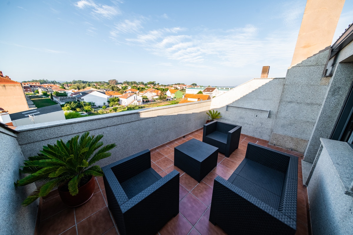A spacious terrace is featured, furnished with four rattan-style seating pieces arranged around a coffee table. A large planter with greenery is positioned in one corner. The view extends over the neighborhood, revealing rooftops and glimpses of the sea in the distance.