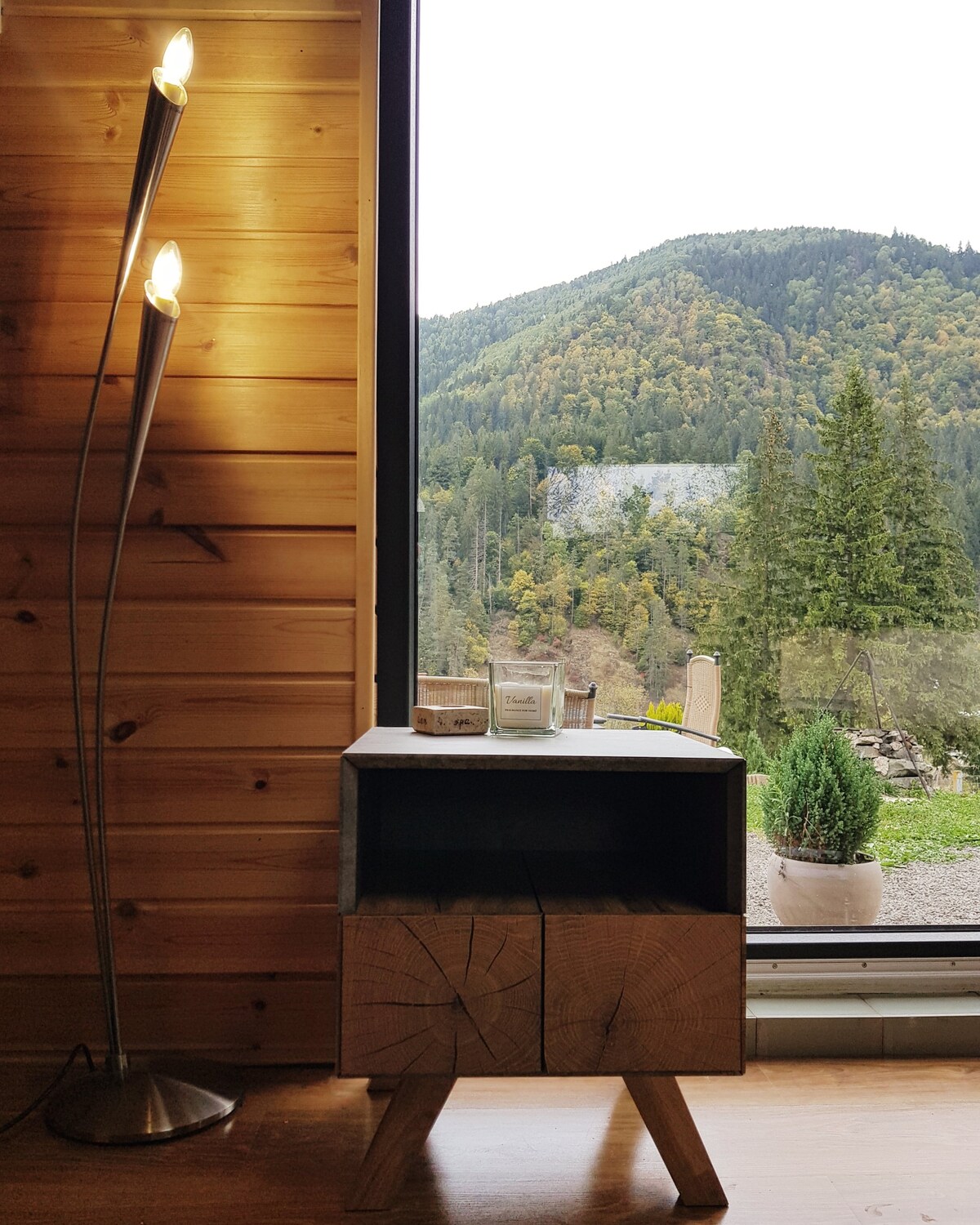 A stylish wooden nightstand with a unique design stands beside a large window. A candle and small items are placed on top, while the window offers a view of lush green hills and trees outside, creating a serene connection with nature.