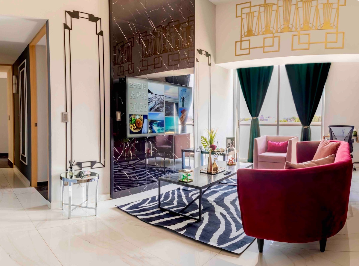 A spacious living area features elegant Art Deco elements, including geometric patterns and high ceilings. A comfortable seating arrangement, comprising a pink armchair and a deep red sofa, is positioned on a striking zebra-patterned rug. Large windows with dark curtains allow ample natural light to fill the room.