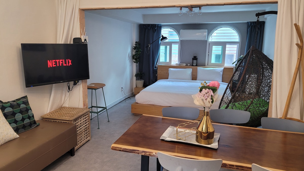 A cozy living area features a television screen displaying Netflix. A dining table is set nearby, accompanied by light-colored chairs. The sleeping area showcases a spacious bed with green accents, and a decorative hanging chair adds a playful touch. Natural light filters through large arched windows.