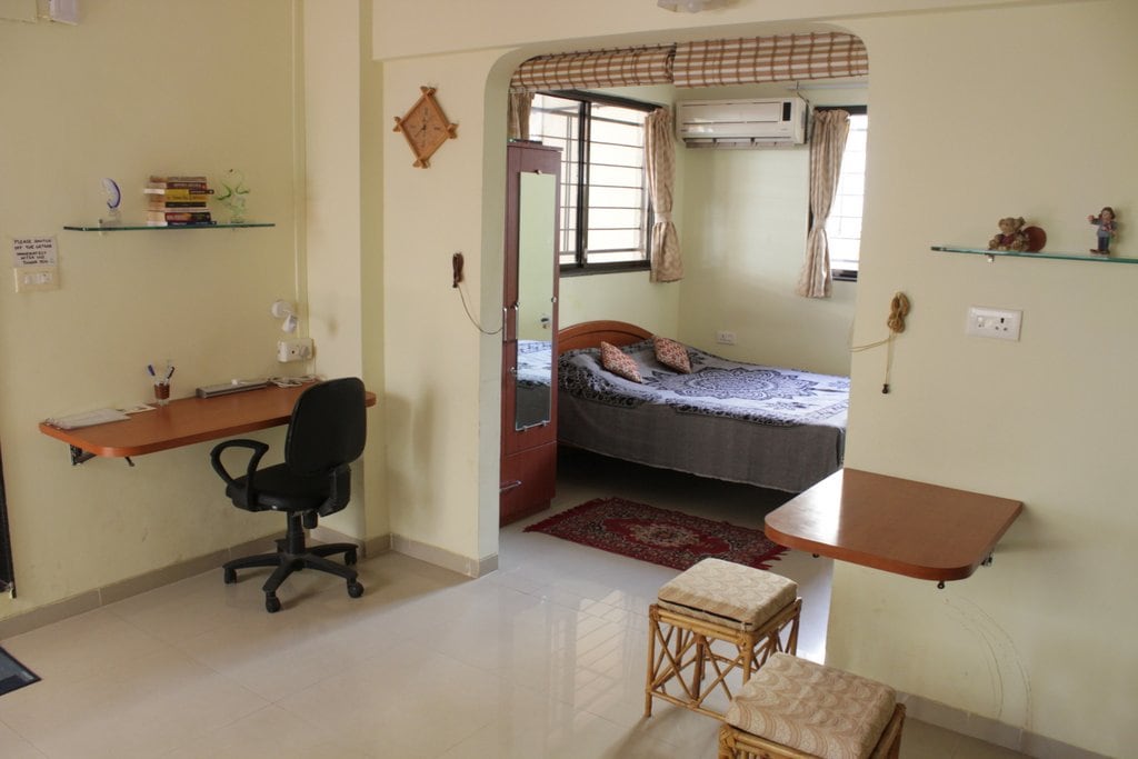 A spacious studio apartment is depicted, featuring a separate sleeping area with a bed partially covered by a patterned blanket. A study desk with a chair is positioned nearby. Natural light enters through large windows, enhancing the simple and functional design of the space.