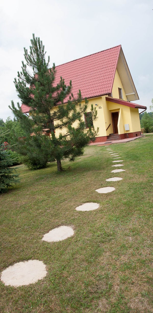 A well-maintained pathway of flat stepping stones leads from the grassy area to a yellow two-story house with a red peaked roof. A young pine tree stands beside the path, enhancing the natural setting.