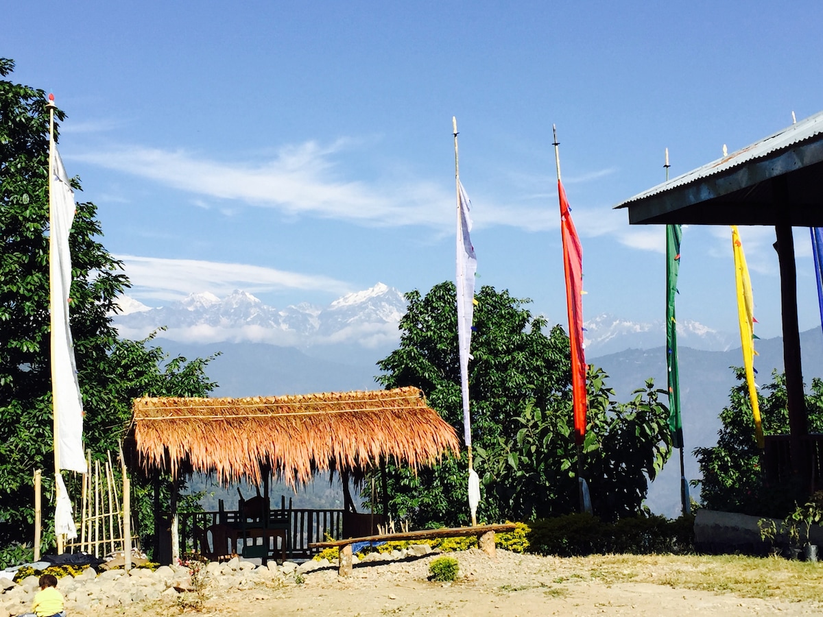 A traditional thatched gazebo is set against a backdrop of snow-capped mountains and clear blue skies. Colorful prayer flags gently wave in the breeze, adding a touch of cultural significance. Lush greenery surrounds the area, enhancing the serene atmosphere.