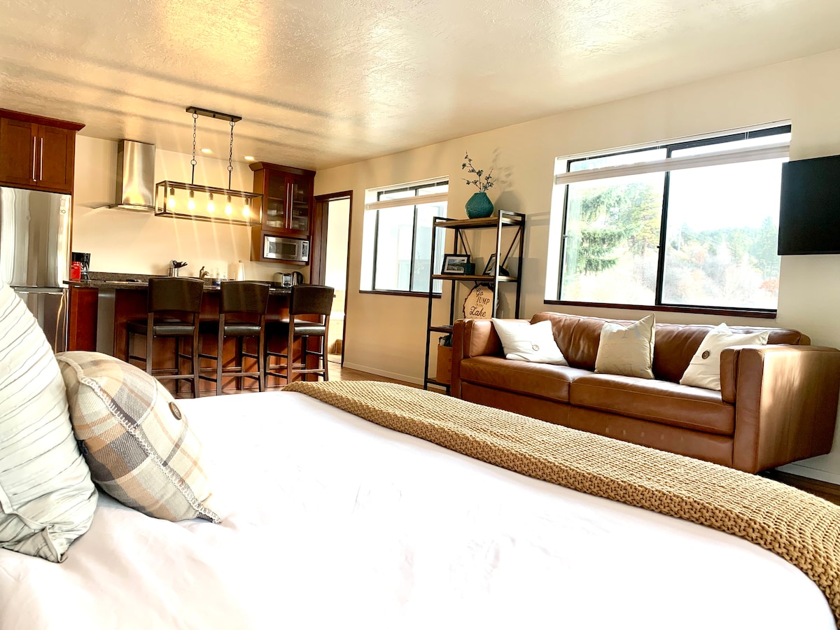 An open and inviting interior showcases a cozy seating area with a brown leather sofa, accented by decorative pillows. The kitchen area features modern appliances and a dining space with four dark wood chairs. Large windows allow natural light to fill the space, enhancing the view.