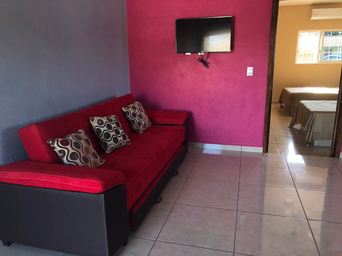 A bright lounge area features a red sofa with decorative pillows, positioned against a purple wall. A flat-screen television is mounted above. Natural light enters from a window leading to a bedroom, which is visible in the background.