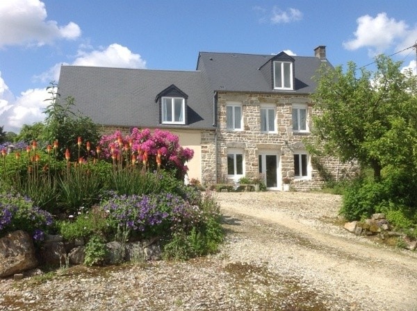 A restored stone farmhouse is set amidst well-maintained gardens featuring vibrant flowers in various hues. The building's gray roof and large windows complement the natural surroundings. A gravel pathway leads to the entrance, surrounded by greenery and seasonal blooms.