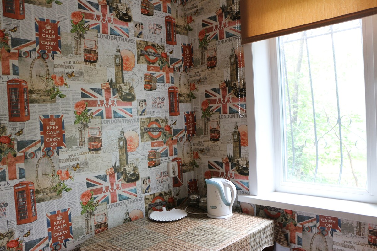 A corner of the kitchen features wallpaper decorated with British motifs, including iconic red phone booths and travel-themed illustrations. A small table is placed beneath a window, which is adorned with a beige roller shade. A white electric kettle rests on the table, alongside a circular plate.