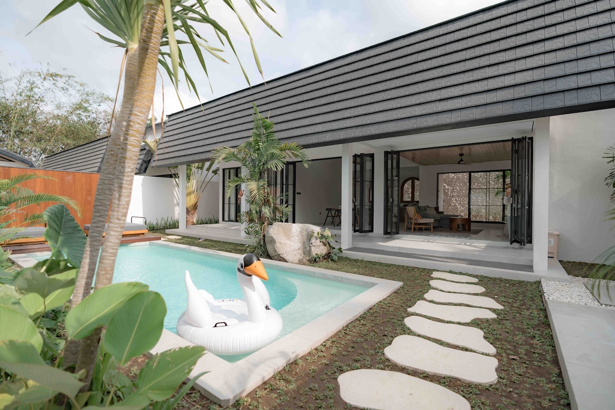 A private pool is featured in the outdoor area, complemented by a floating swan-shaped inflatable. Lush greenery surrounds the pool, while a stone pathway leads toward an open living space with large glass doors, enhancing the connection to the outdoors.