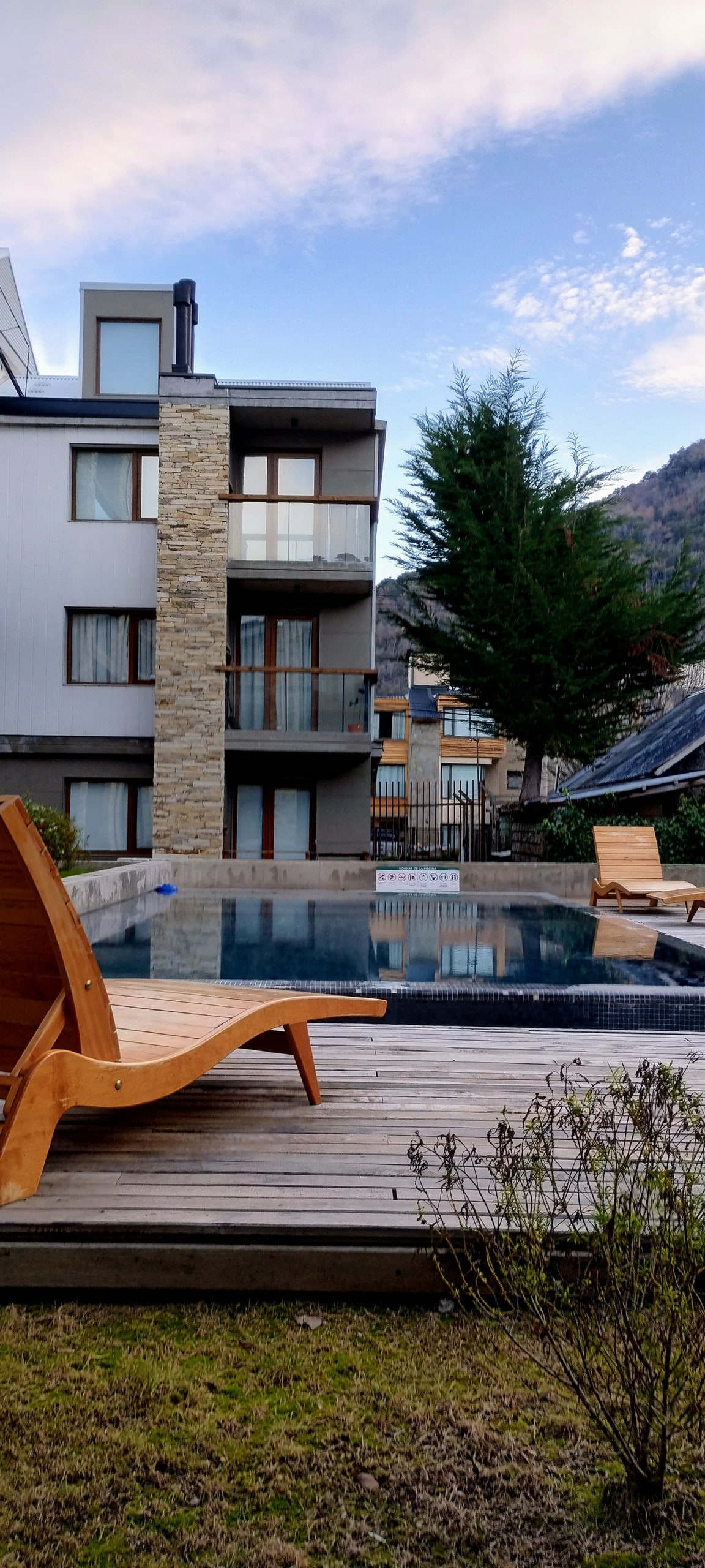 A modern building features a stone facade and glass balconies facing a serene pool area. Wooden lounge chairs surround the pool, which is set against a backdrop of trees and mountains. The well-maintained lawn adds to the inviting outdoor space.