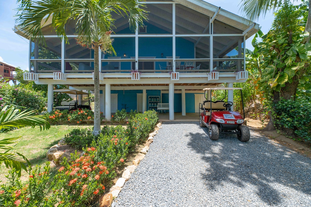 The exterior of the Villa is shown, featuring a spacious screened porch above and a landscaped yard filled with greenery and colorful flowers. A gravel path leads to the entrance, with a golf cart parked nearby for convenient transportation.