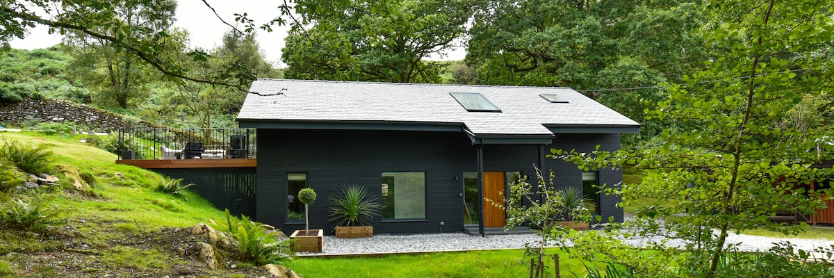 The exterior of a modern, contemporary holiday home is showcased among lush greenery. The building features a combination of dark walls and a grey roof, with large windows and a wooden door. A landscaped area with planter boxes and decorative shrubs is visible.
