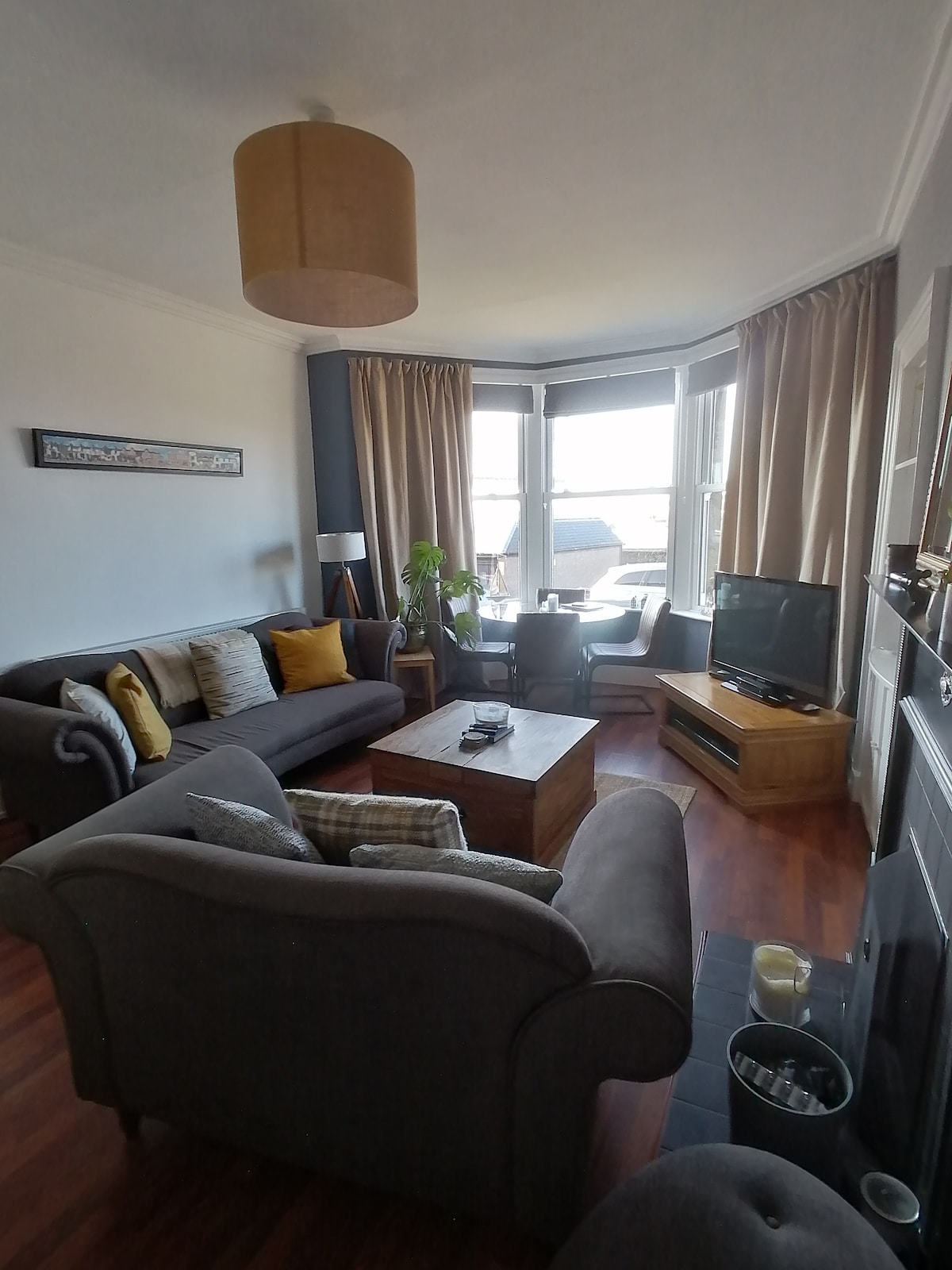 A cozy living area features comfortable seating with plush grey sofas, complemented by decorative cushions. A wooden coffee table sits in the center, surrounded by light streaming through large bay windows with elegant drapes. A flat-screen TV and indoor plants enhance the inviting ambiance.