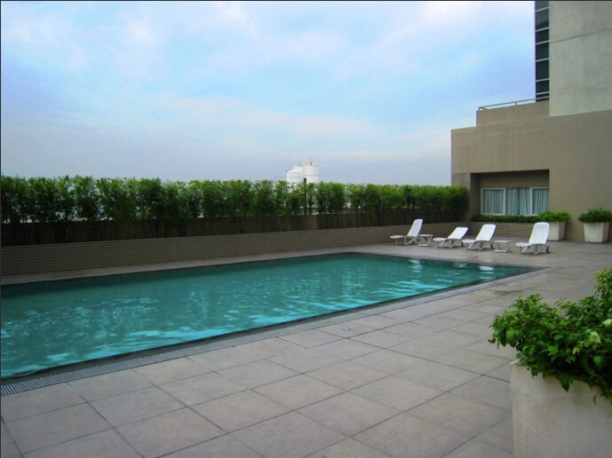 A rectangular pool is set on a large terrace with modern tile flooring. Six lounge chairs are positioned around the pool, offering a place to relax. Lush greenery lines the perimeter, providing a sense of privacy and enhancing the serene atmosphere.