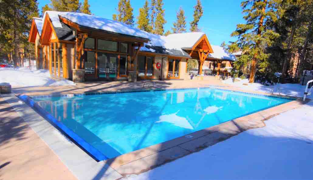 A heated outdoor pool, enveloped by snow-covered pine trees, is visible in the foreground. The pool area includes a wooden pool house with large windows, allowing natural light to fill the space. Lounge chairs surround the pool, providing relaxation options.