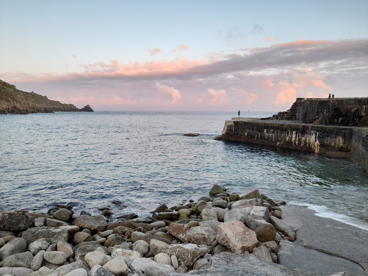A serene coastal scene features calm waters gently lapping against rocky shorelines. Soft pastel hues fill the sky at sunset, with rugged cliffs framing the view. A stone pier extends into the sea, and a few figures can be seen in the distance, enjoying the tranquil atmosphere.