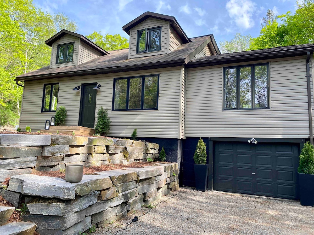 The exterior of the cabin features a two-story structure with large windows, framed by greenery. Stone steps lead to an entrance flanked by plants. The garage door is visible, and the gravel driveway provides ample parking space.