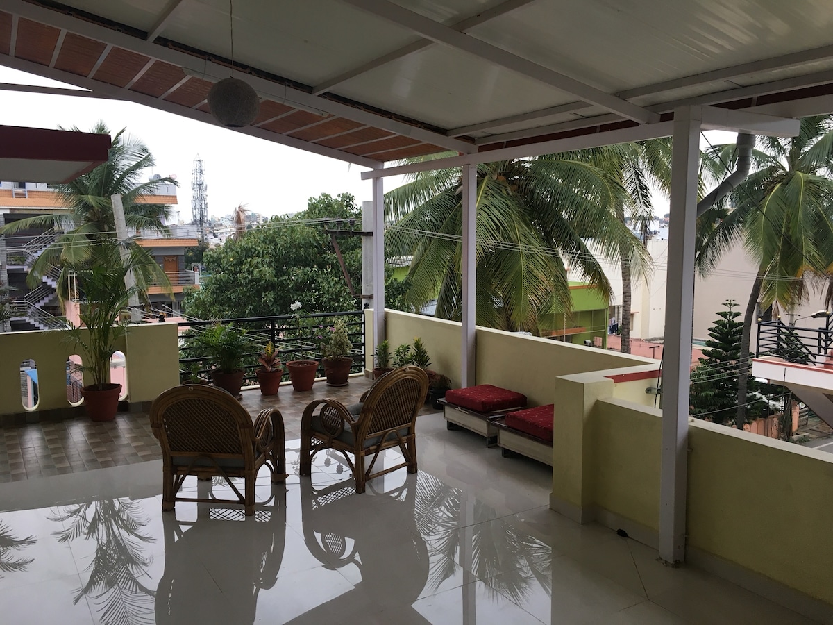 A spacious balcony features comfortable seating made of wicker, with cushions arranged along one side. Lush palm trees are visible beyond the railing, complementing the serene outdoor space. The floor is tiled, reflecting soft natural light that illuminates the area.