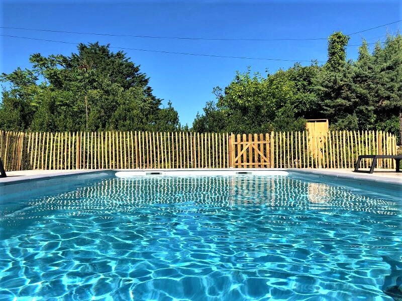 A clear blue swimming pool is visible, surrounded by a wooden fence. Lush greenery is seen in the background, providing a natural setting. The bright blue sky above enhances the tranquil atmosphere of the outdoor space.