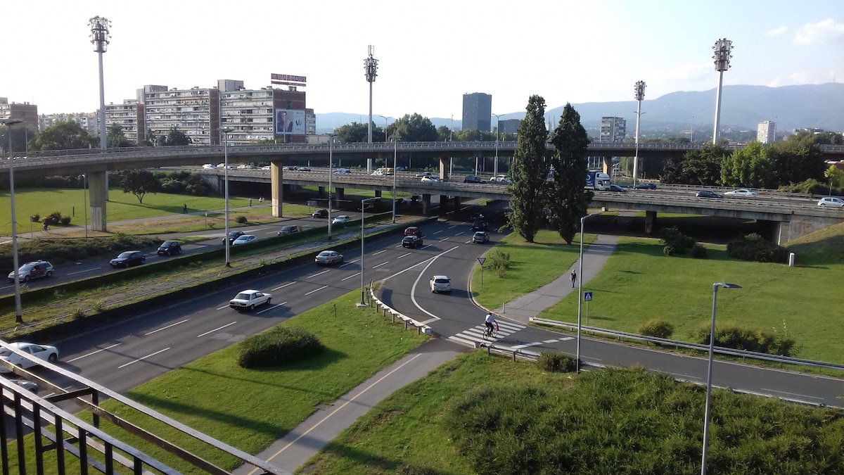 A view of the surrounding area features a busy roadway with multiple lanes and vehicles in motion. Lush green grass and trees are visible along the edges, while distant buildings and mountains provide a scenic backdrop. Streetlights and sports field lights are positioned throughout the scene.