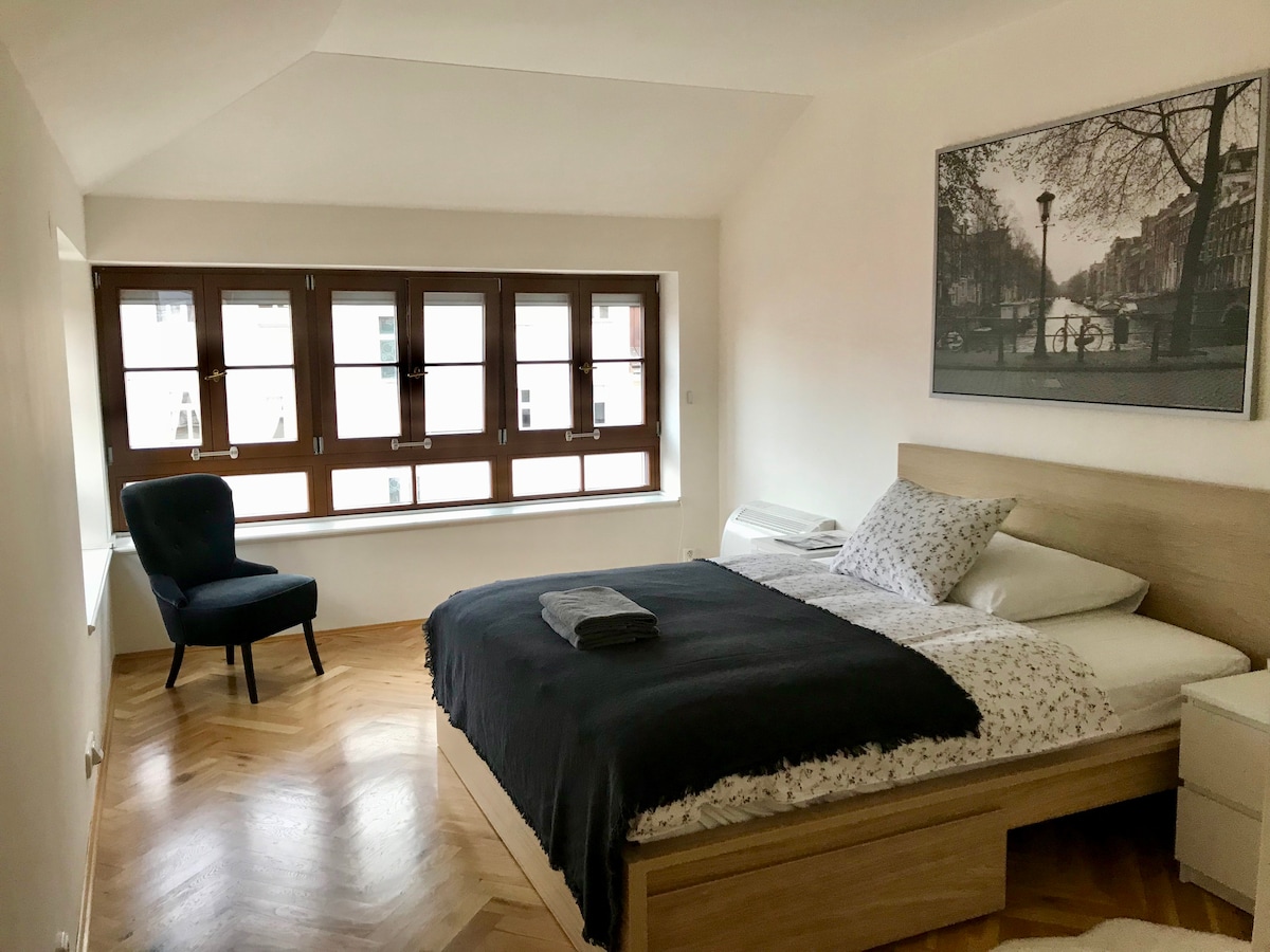 The bedroom features a spacious bed dressed in neutral tones, complemented by a large black throw. A cozy chair sits by the five-window arrangement, which allows ample natural light to fill the room. Light wood flooring enhances the airy atmosphere.
