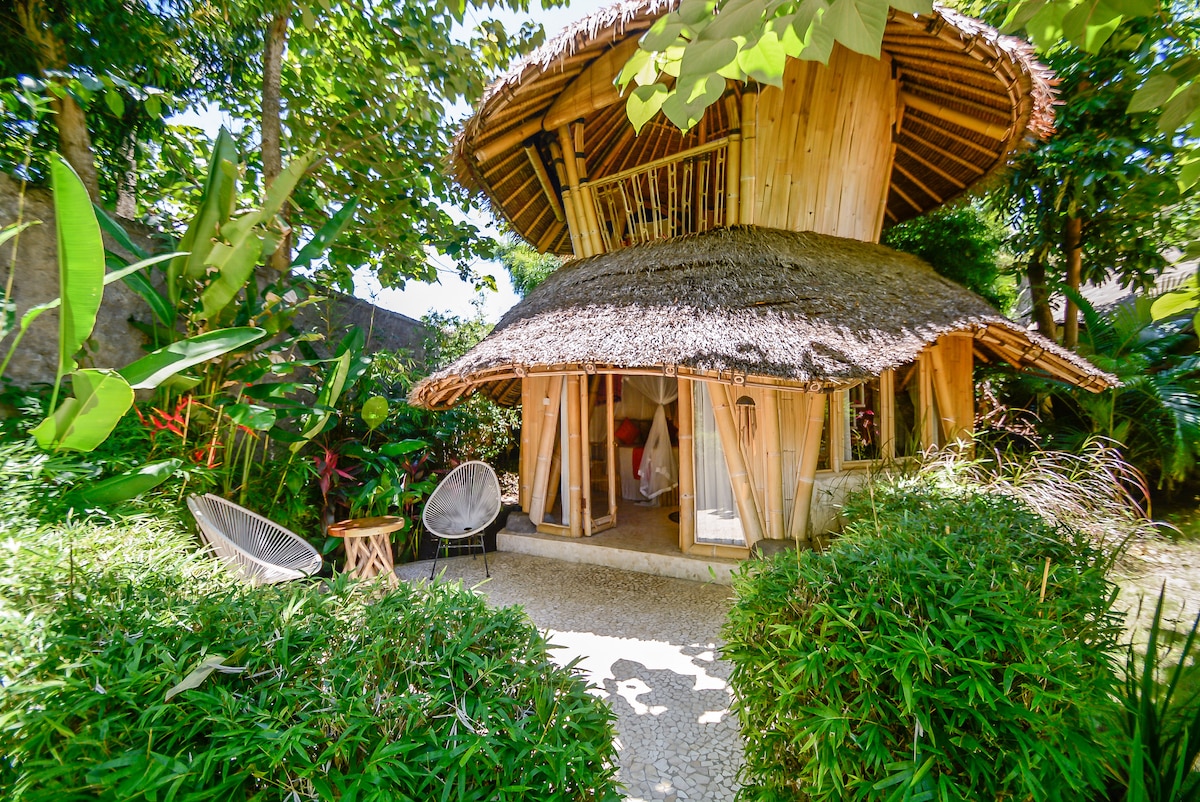 A uniquely crafted two-story bungalow is set amidst lush greenery, showcasing natural materials and a thatched roof. The entrance is framed by vibrant plants, and a small seating area with two chairs is positioned on the stone pathway leading to the door.