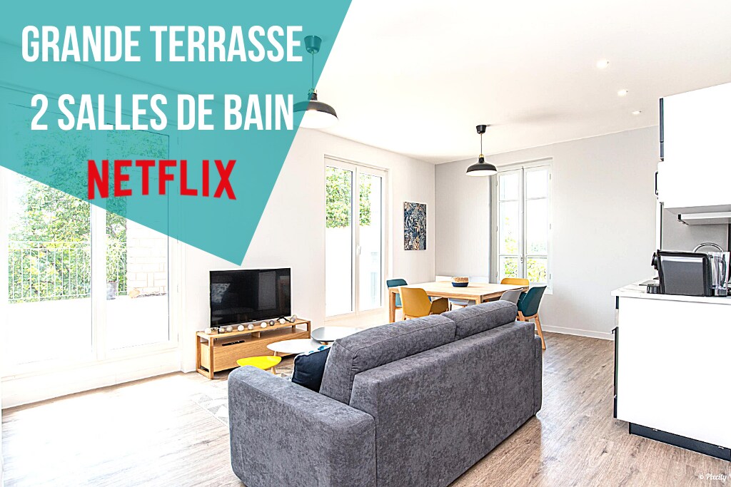 A spacious living area features a comfortable grey sofa, complemented by bright yellow and blue chairs. Large windows allow natural light to fill the room, offering views of the terrace. The open layout connects to a modern kitchen, enhancing the inviting ambiance.
