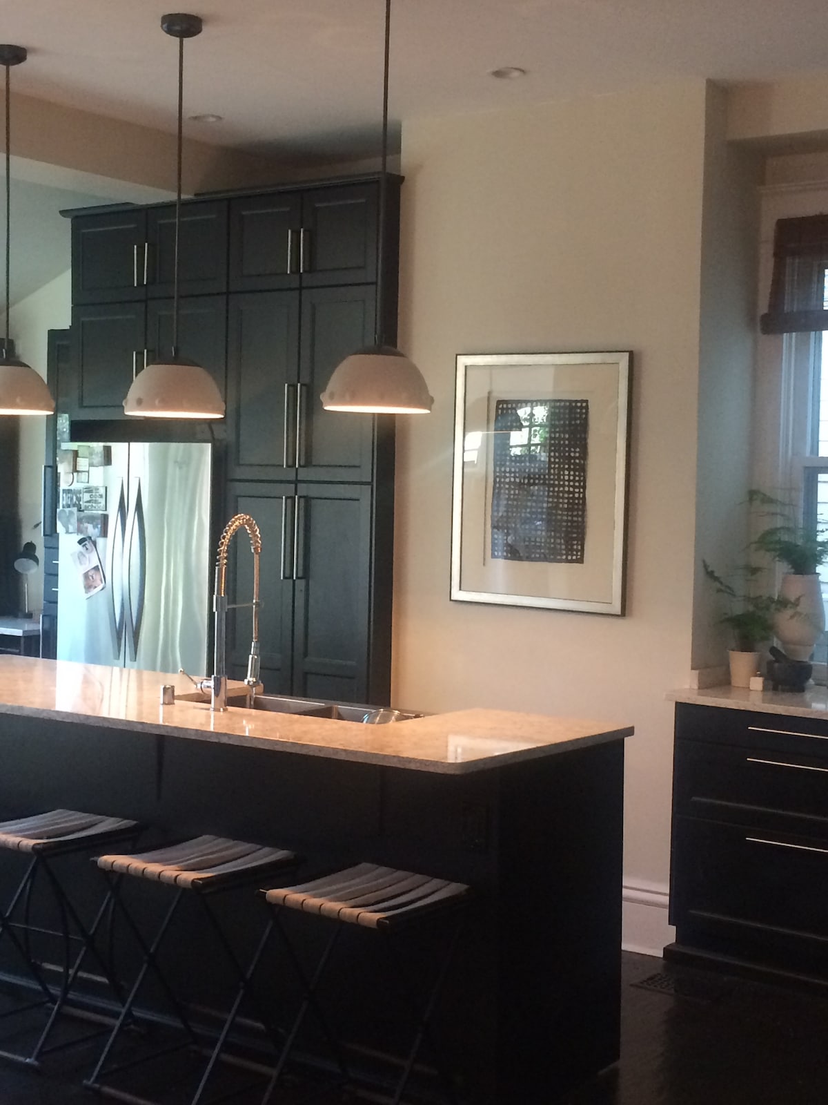 A sleek kitchen is shown, featuring dark cabinetry and a light granite countertop. Three stools with striped cushions are positioned at the kitchen island. Warm lighting is provided by pendant fixtures above. A framed artwork adds a touch of decor, and a plant is visible on the counter.