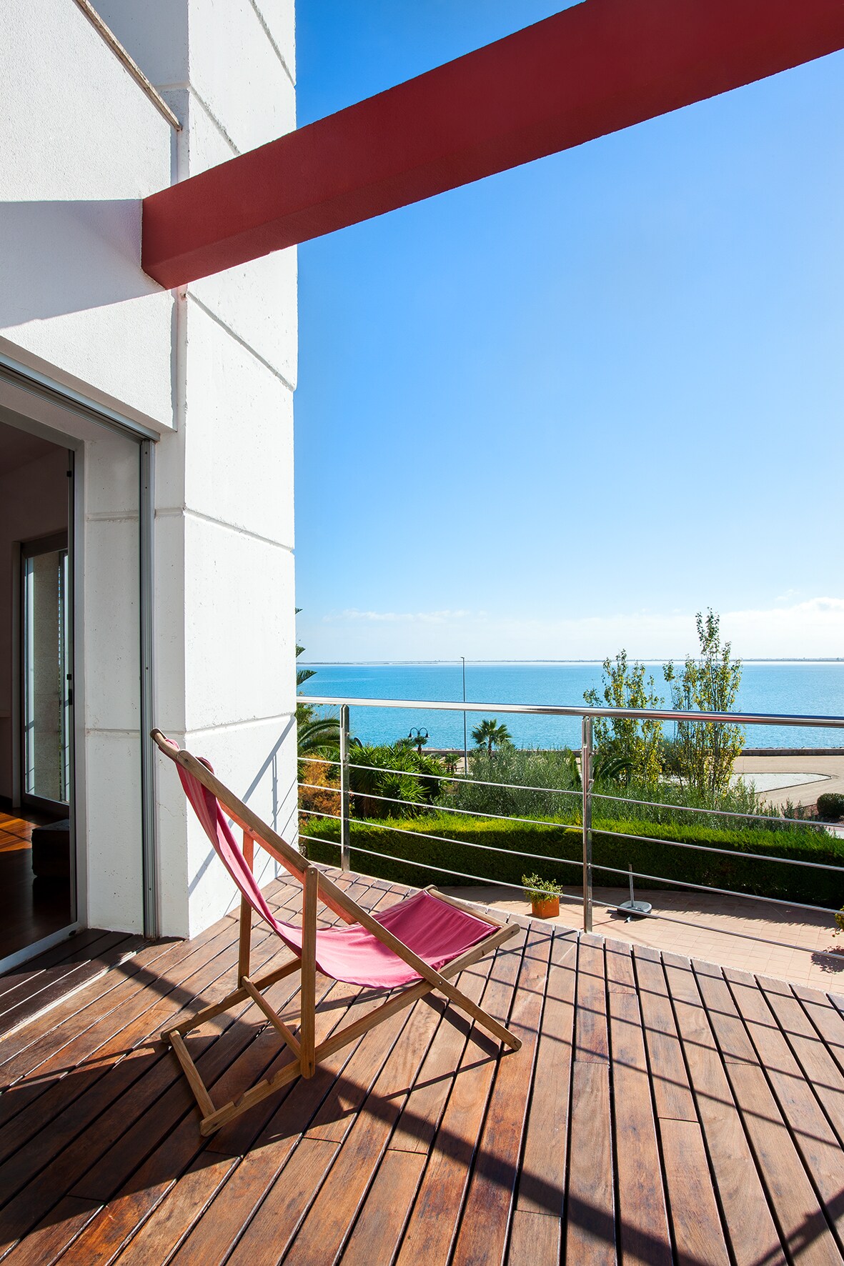 A wooden terrace extends from the villa, showcasing a folding chair facing the sea. Bright sky and sea views create a serene backdrop, while greenery is visible in the distance, enhancing the outdoor atmosphere.