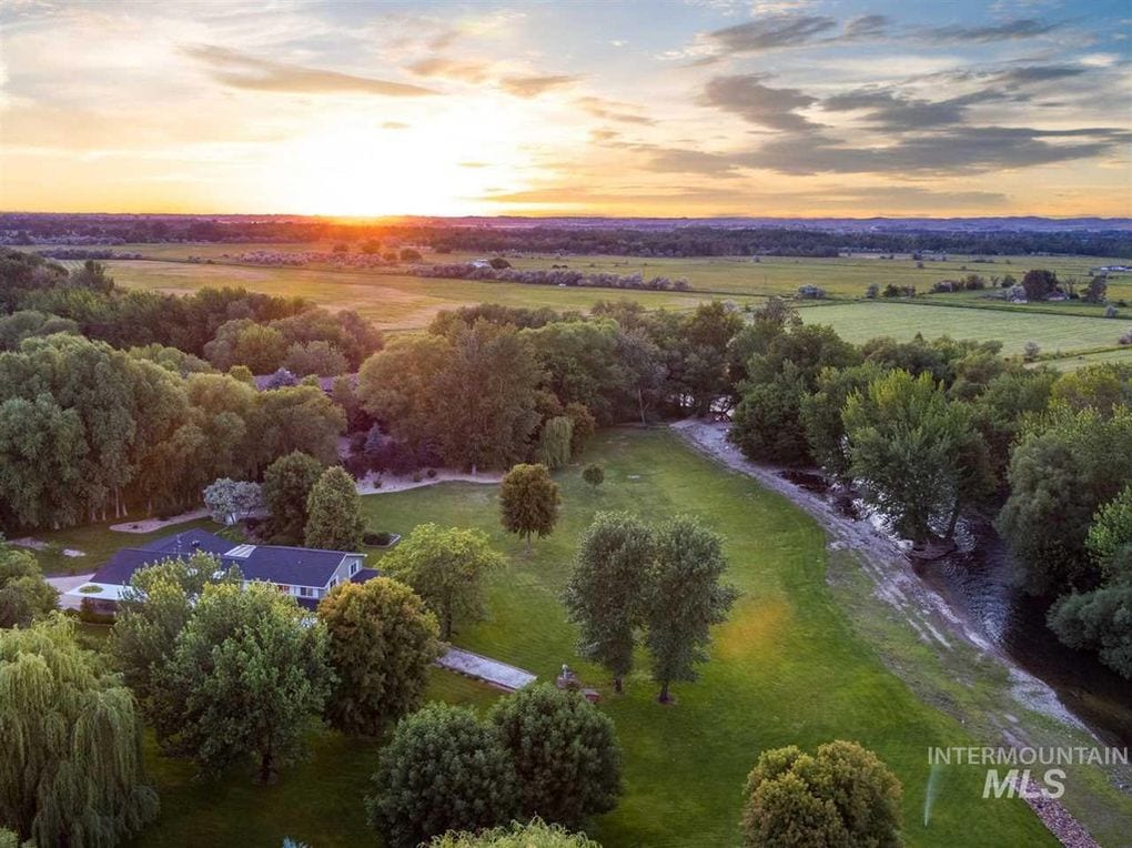 Aerial view captures the vast green lawn and mature trees surrounding the property, contrasted by the winding Boise River. The sun sets in the background, casting warm hues across the landscape, highlighting the beauty of the natural setting.