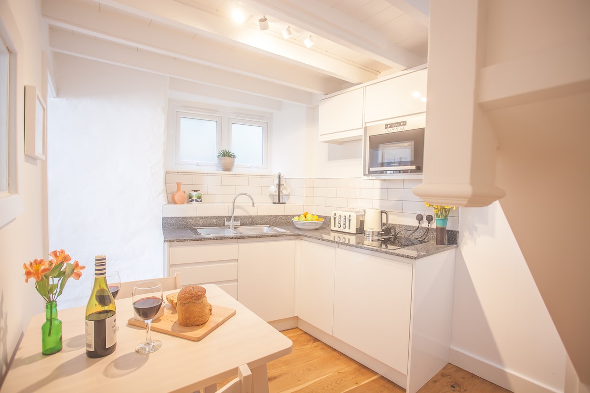 The modern kitchen is equipped with sleek white cabinetry and granite countertops. A small dining table is set for two, featuring a bottle of wine and bread. Natural light floods the space through two small windows, enhancing the clean and functional design.
