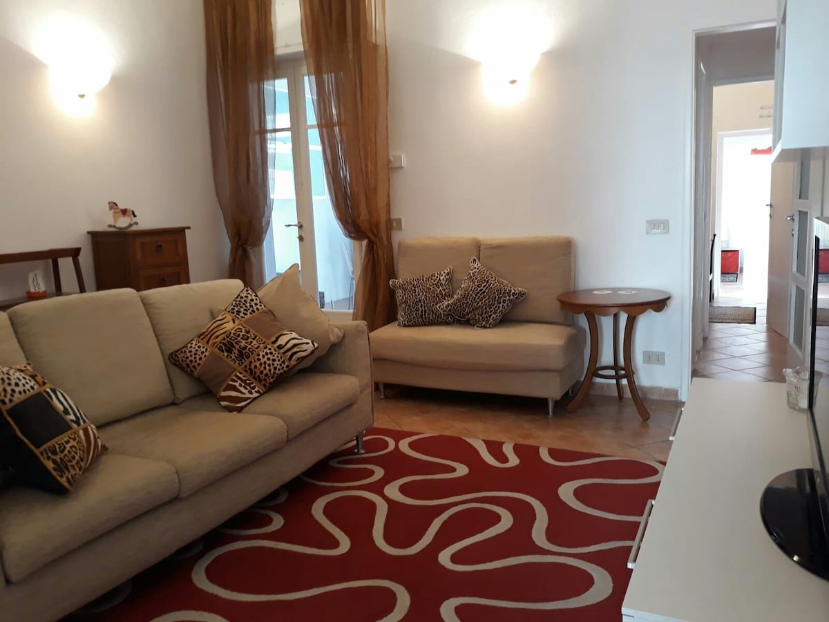 A living area is presented with a neutral-colored sofa and a compact loveseat, accompanied by decorative pillows featuring animal prints. A round table is positioned beside the loveseat. Soft lighting is provided by wall sconces, with a patterned area rug adding color to the tiled floor.