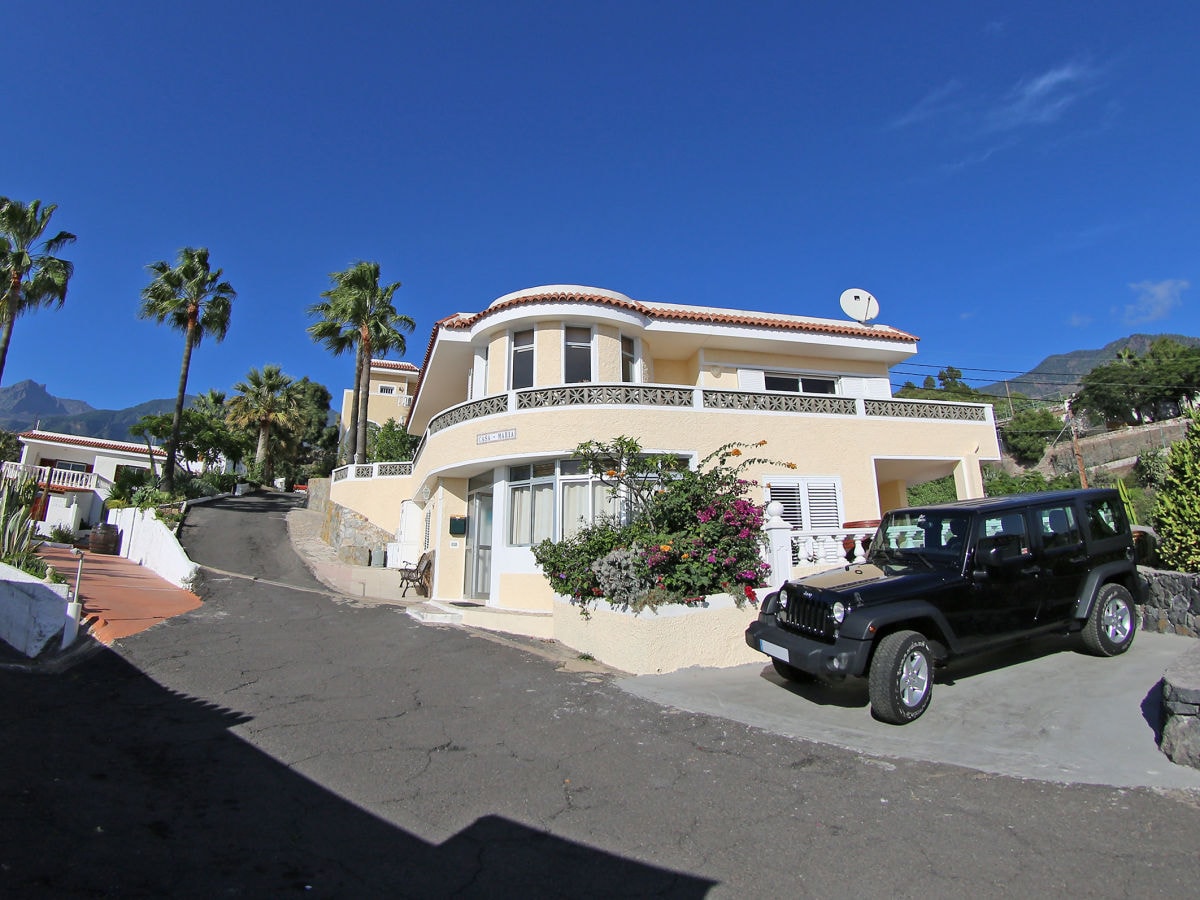 The exterior of the Casa Maria features a multi-level structure framed by tropical palm trees. A black vehicle is parked on the driveway, while the entrance is adorned with colorful plants. Clear blue skies complement the warm hues of the building.