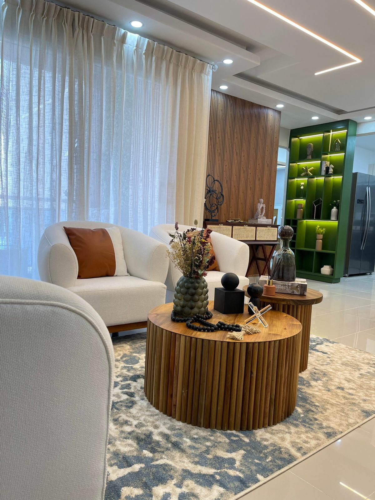 A modern living area is showcased with two upholstered armchairs positioned beside a round wooden coffee table. Decorative items, including a succulent plant and candles, are arranged on the table. Natural light filters through sheer curtains, illuminating the space with a warm ambiance.