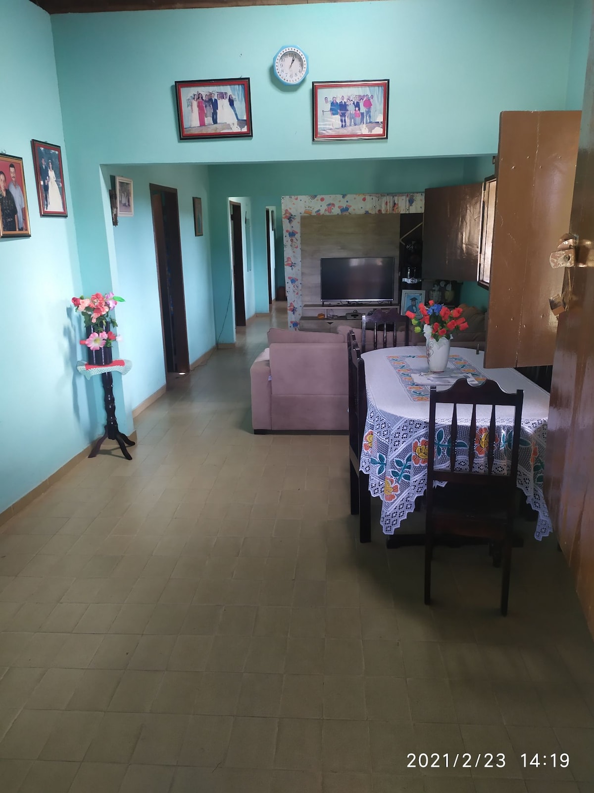 A hallway connects the living and dining areas, showcasing a light green color palette. Family photographs adorn the walls. A table with a decorative tablecloth is set for dining, accompanied by chairs. A cozy sofa is positioned near a television in the adjacent living area.