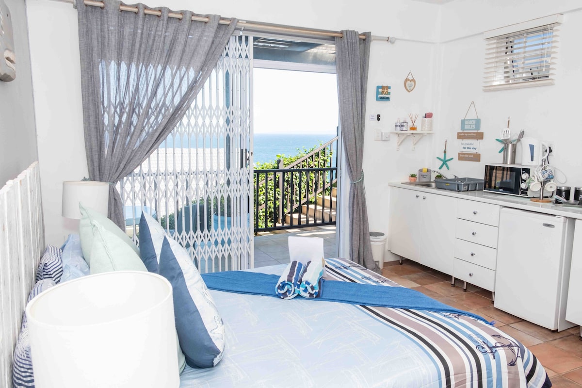 A bright studio cottage features a spacious bed adorned with blue and white linens. Natural light floods the space through a glass door leading to a covered veranda with ocean views. A small kitchenette with essential appliances, including a microwave and coffee maker, is visible along the wall.